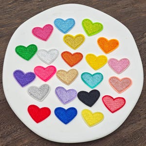 Peut inclure: Une collection de vingt-cinq petits écussons brodés en forme de cœur dans une variété de couleurs, notamment rouge, rose, orange, jaune, vert, bleu, violet, noir, argent et or, disposés sur une surface blanche circulaire.
