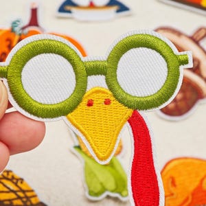 Puede incluir: Un parche bordado que muestra la cara de un pavo con gafas redondas de color verde brillante, un pico amarillo con dos pequeñas fosas nasales rojas y una larga carúncula roja colgante, todo bordeado con hilo blanco sobre fondo claro.