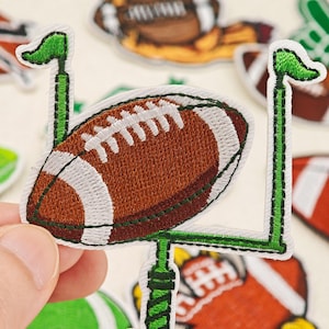 Könnte beinhalten: Ein gestickter Aufnäher mit einem braunen American Football mit weißen Schnürsenkeln vor einem grünen Torpfosten. Der Aufnäher hat einen weißen Rand und wird vor einem Hintergrund aus weiteren ähnlichen, football-thematischen Aufnähern gehalten.