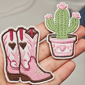 Peut inclure: Deux écussons brodés représentant une paire de bottes de cowboy blanches et roses avec des cœurs marron et un cactus vert dans un pot rose avec des fleurs roses. Les écussons ont des bordures blanches et des coutures détaillées.