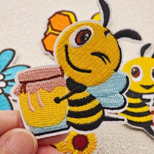 Peut inclure: Un écusson brodé thermocollant représentant une abeille de dessin animé au corps rayé jaune et noir, aux ailes bleu clair et au visage souriant, tenant un petit pot de miel doré avec un couvercle rose, entouré d'autres écussons floraux.