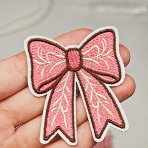 Peut inclure: Un écusson brodé rose en forme de nœud. Le nœud présente un contour cousu marron foncé et un nœud central, avec des motifs de vigne crème clair brodés sur la surface des sections en tissu rose de l'accessoire textile.
