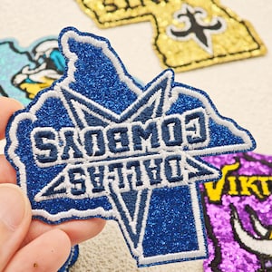 Peut inclure: Un écusson brodé bleu à paillettes en forme de l'État du Texas, arborant une étoile blanche et le texte Dallas Cowboys brodé en bleu foncé. L'écusson présente une bordure blanche cousue et est présenté devant d'autres écussons sportifs.