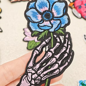 Op de afbeelding: Een geborduurde patch met een skeletachtige hand in lichtroze en witte tinten, die een levendige blauwe bloem met een donker hart en groene bladeren vasthoudt. Het ontwerp is omlijnd met zwart garen voor een contrastrijke grafische uitstraling.