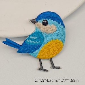 Peut inclure: Un écusson brodé représentant un petit oiseau avec une tête et un dos bleu vif, une face beige clair, un ventre jaune et des pattes gris foncé. L'oiseau a un œil noir et un petit bec. L'écusson mesure 4,5 cm sur 4,2 cm comme indiqué.