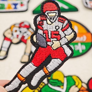 May include: An embroidered fabric patch featuring a football player in a white and red uniform with the number 15 on the chest, wearing a red helmet and holding a dark football, outlined in black thread against a light background.