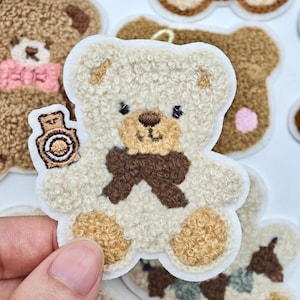 May include: A cream-colored teddy bear patch with a textured, fuzzy surface. The bear features a dark brown bow tie, a brown nose, and black eyes. It holds a small brown and white object in its right paw. The patch has a white border.