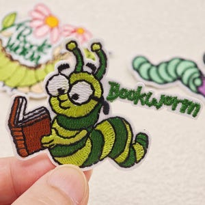 Peut inclure: Un écusson brodé représentant un ver de terre vert de dessin animé avec de grands yeux blancs, portant des lunettes et tenant un livre brun ouvert. Le mot Bookworm est brodé en fil vert à droite du personnage, sur un support en tissu blanc.