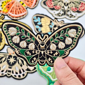 Può includere: Una toppa ricamata a forma di falena su base in tessuto nero con dettagli in filo dorato e verde. Il design presenta accenti floreali bianchi sulle ali e un simbolo centrale a mezzaluna. Altre toppe sono visibili sullo sfondo.