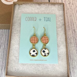May include: A pair of gold dangle earrings with two circular charms. The top charm is a woven peach-colored fabric and the bottom charm is a white fabric with black polka dots. The earrings are displayed on a blue card with the text "Copper + Teal" and the website address "www.copperandteal.com".