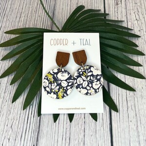 Puede incluir: Un par de pendientes de madera con un patrón floral en la parte delantera. Los pendientes son redondos y tienen un fondo azul oscuro con flores blancas y amarillas. Los pendientes cuelgan de un poste de madera con un acabado marrón. Los pendientes están en una tarjeta blanca con el texto "Copper + Teal" y "www.copperandteal.com".