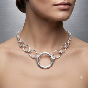 May include: A silver-toned necklace featuring a series of interconnected circular links that gradually increase in size toward the center. The central element is a larger, textured circular pendant, with smaller rings extending outward to form the chain.