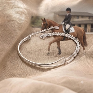 May include: A silver-toned bracelet featuring a smooth, curved metal bar connected to a textured, sparkling chain segment. The piece includes a delicate extender chain with a small, flat, teardrop-shaped charm and a secure circular spring clasp.