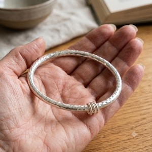 Może przedstawiać: Srebrzysta bransoletka typu bangle o młotkowanej, teksturowanej powierzchni. Okrągła bransoletka posiada mały, gładki pierścionek w kolorze srebrnym, który swobodnie przesuwa się wzdłuż głównej obręczy, tworząc prosty metalowy dodatek.