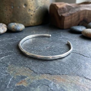 May include: A silver cuff bracelet with a smooth, polished finish and an open-ended design. The metal band features a slightly irregular, organic shape with subtle indentations along its surface, creating a reflective and bright metallic appearance.