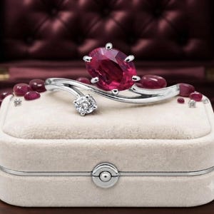 Puede incluir: Un anillo bypass de tono plateado con un gran rubí rojo oscuro de corte ovalado y un diamante blanco transparente más pequeño de corte redondo. El anillo se exhibe sobre un joyero rectangular de terciopelo beige, rodeado de pequeños rubíes rojos pulidos.