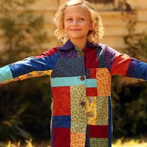 Puede incluir: Un abrigo de manga larga con un diseño de patchwork vibrante compuesto por varias piezas de tela rectangulares en tonos rojo, azul, amarillo y turquesa. La prenda incluye un cuello morado sólido y tres botones marrones en la parte delantera.