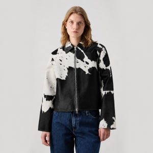 May include: A long-sleeve jacket featuring a black and white cow print pattern. The jacket has a classic collar, a full-length silver-toned metal zipper closure down the center, and a straight hemline. The material has a textured, hair-on-hide appearance.