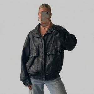 May include: A black leather jacket featuring a collared neckline, long sleeves with buttoned cuffs, and two front pockets with snap closures. The jacket has a front zipper closure and a ribbed hemline, paired with light blue denim jeans.