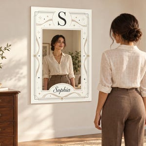 Puede incluir: Un espejo de pared rectangular con un borde decorativo que presenta la letra S en la parte superior y el nombre Sophia en letra cursiva en la parte inferior. El marco incluye líneas negras finas, formas geométricas y estrellas sobre fondo blanco.