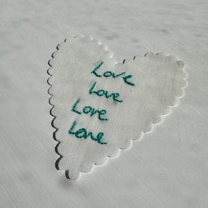 May include: A white fabric heart with scalloped edges featuring the word Love embroidered four times in teal thread. The embroidery is stacked vertically in the center of the heart, showcasing a hand-stitched appearance on the white textile.