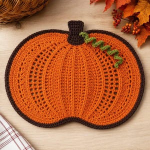 May include: A pumpkin-shaped crochet placemat featuring a vibrant orange center with a textured stitch pattern, bordered by a dark brown edge. It includes a dark brown stem at the top and a single, spiraled green vine accent attached to the side.