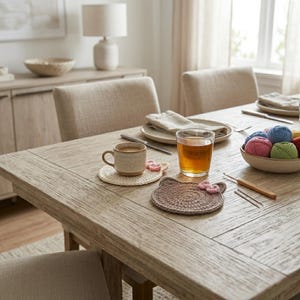 May include: Two round crochet coasters, one cream and one brown, featuring small pink flower accents. The brown coaster is shaped like a cat head with ears. A ceramic mug sits on the cream coaster, and a wooden crochet hook lies nearby.