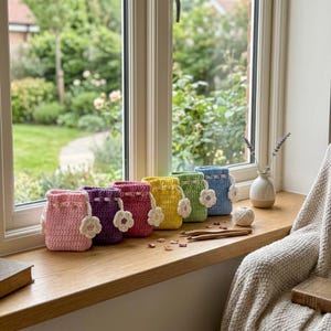 May include: A row of six small, crocheted drawstring pouches in pastel pink, purple, magenta, yellow, green, and blue. Each pouch features a white crocheted flower attached to the drawstring cord. The pouches are arranged on a wooden ledge.