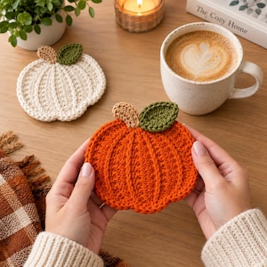 Könnte beinhalten: Zwei runde, kürbisförmige Häkeluntersetzer. Ein Untersetzer ist leuchtend orange, der andere cremeweiß. Beide haben einen kleinen, hellbraunen Stiel und ein einzelnes dunkelgrünes Blatt an der Oberseite. Die Textur ist gerippt und kreisförmig.