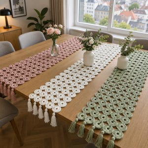 May include: Three rectangular crochet table runners in dusty pink, white, and sage green, each featuring a repeating circular pattern and finished with matching fringe tassels at the ends, displayed on a wooden dining table with flower vases.