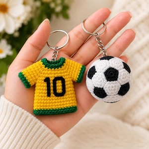 Puede incluir: Dos llaveros de ganchillo que incluyen una camiseta amarilla con bordes verdes y el número 10 negro en la parte delantera, y un balón de fútbol esférico con un patrón geométrico clásico en blanco y negro. Unidos a anillas de metal plateado.