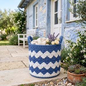 May include: A round, crocheted storage basket featuring a repeating chevron pattern in alternating bands of deep blue and white yarn. The basket has two integrated side handles and contains a light-colored fabric liner with lavender and roses.