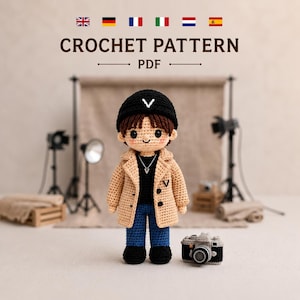 Op de afbeelding: Een gehaakte pop met een zwarte muts met een witte V, een beige jas met knopen, een zwart shirt en een blauwe broek. De pop heeft bruin haar en een kleine ketting. Een kleine gehaakte camera staat ernaast. Tekst leest CROCHET PATTERN PDF.