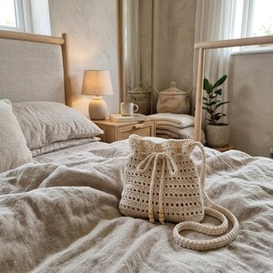 May include: A cream-coloured crochet drawstring bag with a long shoulder strap. The bag features an open-weave pattern, a cinched top with two matching cord ties, and two small wooden beads at the end of each drawstring cord for a decorative finish.