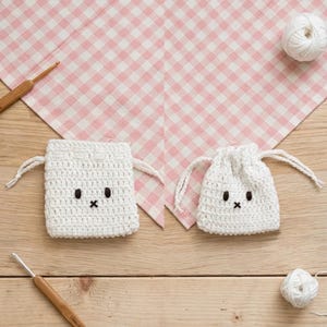 May include: Two white crocheted drawstring pouches featuring embroidered black eyes and a small black cross-stitched mouth. The pouches are accompanied by two wooden crochet hooks and two small balls of white yarn, arranged on a light wood surface.
