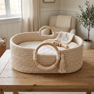 Può includere: Un cesto ovale all'uncinetto color crema con due manici circolari in legno avvolti da filo coordinato. All'interno sono visibili un materassino bianco trapuntato, un panno beige piegato e un piccolo sonaglio in legno. Una nappa decorativa in filo pende da uno dei manici.