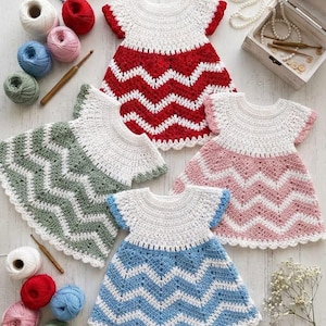 May include: Four handmade crochet dresses for infants, each featuring a white yoke and a chevron-patterned skirt in red, sage green, light pink, or light blue. The set includes various yarn balls, a wooden crochet hook, and a small wooden box.
