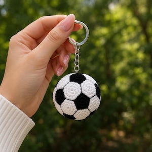 May include: A spherical crochet soccer ball keychain featuring a classic black and white pentagonal and hexagonal pattern. The soft, yarn-based ball is attached to a silver-toned metal split ring by a short, sturdy metal chain link connector.