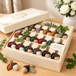 May include: A collection of crocheted acorn keychains with attached fabric leaves in various colours, arranged in a white gift box. Each keychain features a tan crocheted acorn cap and a cream-coloured base. A tag reads Handcrafted Forest Treasures.