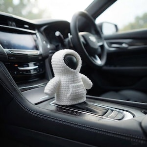 May include: A small, white, crocheted hoodie cover designed to fit over a car gear shift knob. The textile accessory features a hood, long sleeves, and a front kangaroo pocket, crafted with a consistent, textured stitch pattern throughout the piece.