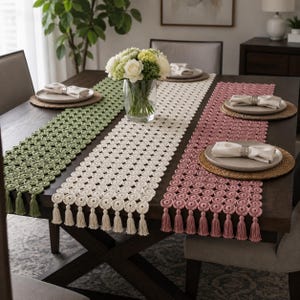 May include: Three rectangular crochet table runners in sage green, cream, and dusty rose, each featuring a repeating circular pattern and finished with matching tassels on the ends. The runners are displayed on a dark wood table with plates.