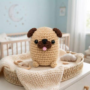 May include: A tan crochet pug plush toy with dark brown ears, a dark brown muzzle, black plastic eyes, and a small pink tongue. The stuffed animal has a rounded body and sits on a cream-colored knitted blanket inside a woven basket.