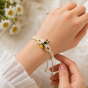 May include: A beige macrame bracelet featuring a small, crocheted bumblebee charm in yellow, black, and white thread. The bracelet is constructed with a knotted cord design and includes adjustable pull strings at the end for a secure fit.