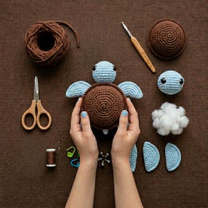 May include: A crochet turtle kit featuring a brown and light blue yarn turtle body, a ball of brown yarn, a wooden crochet hook, metal scissors, a spool of thread, a needle, plastic stitch markers, stuffing, and additional crochet parts.