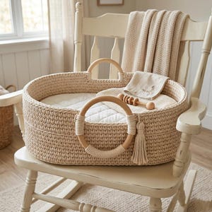 May include: An oval-shaped beige crochet baby bassinet with two circular wooden handles and a matching tassel. Inside, a white quilted mattress pad, a folded beige cloth, and a small wooden rattle are placed. The bassinet sits on a wooden chair.
