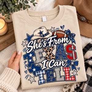 Op de afbeelding: Een beige t-shirt met een grafische print van een cowboyhoed, een football en de tekst "She's From TEXAS I Can TELL". Het ontwerp bevat blauwe, rode en zilveren accenten met luipaard-, paisley- en ruitpatronen, aangevuld met sterren en strikken.