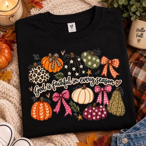 Op de afbeelding: Een zwart t-shirt met een grafische print van diverse pompoenen, strikken en bladeren. De pompoenen zijn versierd met luipaardprint, bloemmotieven en strepen. De tekst luidt, God is faithful in every season, in een cursief handschrift.