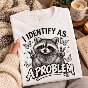 Puede incluir: Una camiseta blanca con una ilustración en tinta negra de un mapache haciendo signos de paz con sus patas. El texto "I IDENTIFY AS A PROBLEM" está impreso en letras negras en negrita, rodeado de pequeños detalles de mariposas y estrellas.