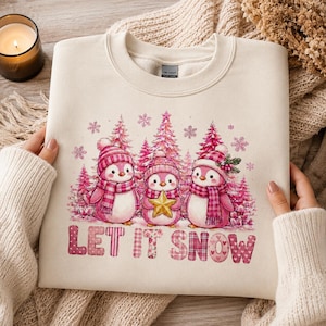 Op de afbeelding: Een crèmekleurige sweater met ronde hals en een afbeelding van drie roze pinguïns met geruite mutsen en sjaals in een roze bos. De middelste pinguïn houdt een gouden ster vast. Onder de afbeelding staat de tekst LET IT SNOW in roze letters.