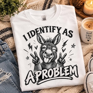 Peut inclure: Un t-shirt blanc à manches courtes avec un motif noir représentant un âne qui fait un clin d'œil et le signe de la paix avec ses sabots. Le texte indique I IDENTIFY AS A PROBLEM, entouré de petits éclairs, cœurs et étoiles dessinés.